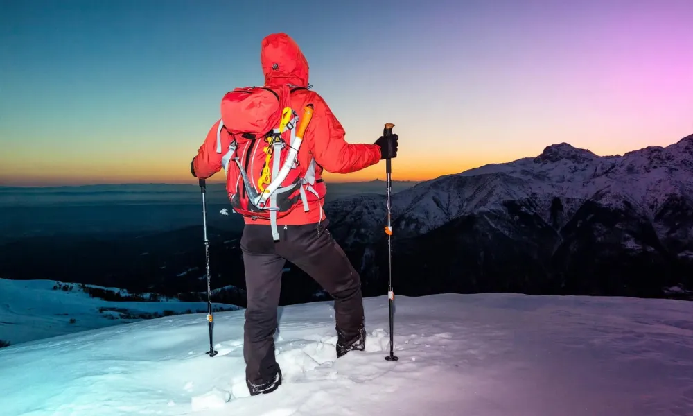 A mountaineer in winter gear holding trekking poles on a snowy summit at sunset-Real Life Inspiring Stories