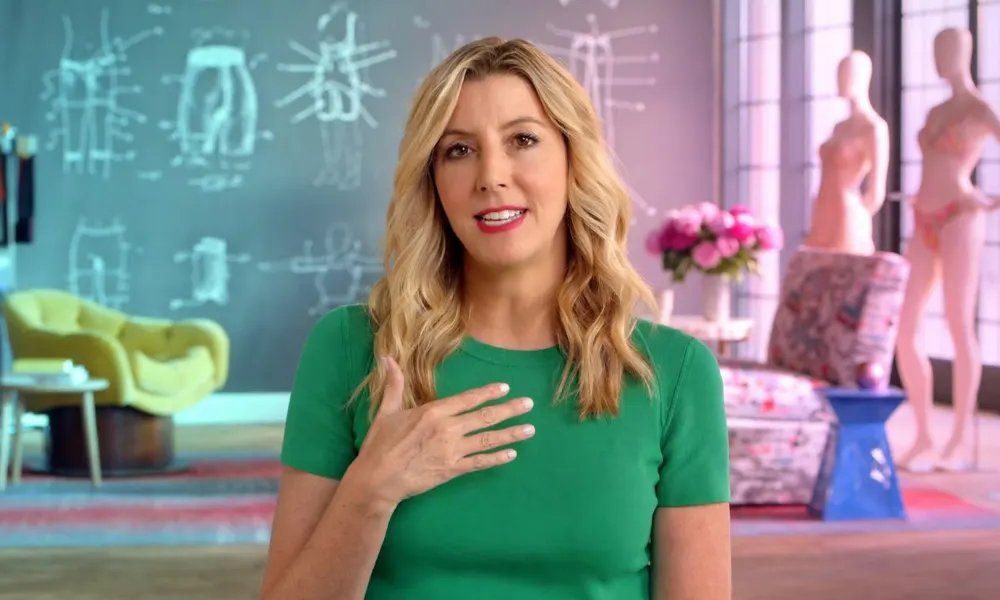 Sara Blakely speaking in a studio with design sketches on a chalkboard behind her.