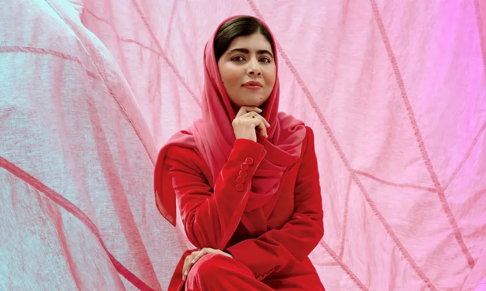 Malala Yousafzai wearing a red suit and headscarf while seated against a pink backdrop-Real Life Inspiring Stories