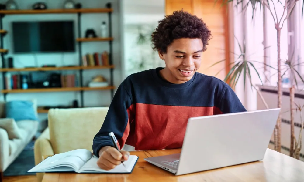 A young student working on a laptop and writing notes at a desk in a living room.