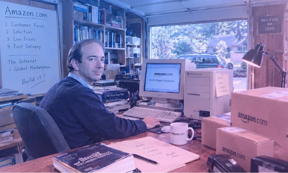 Facezem | How Did Jeff Bezos Start Amazon? The Story Behind Its Beginning Jeff Bezos seated at a desk inside a Bellevue garage workspace surrounded by computer equipment and Amazon shipping boxes