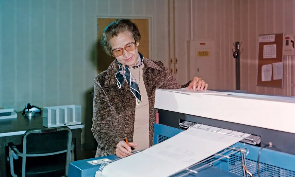 Katherine Johnson reviewing computer printout data at a desk inside a NASA facility.
