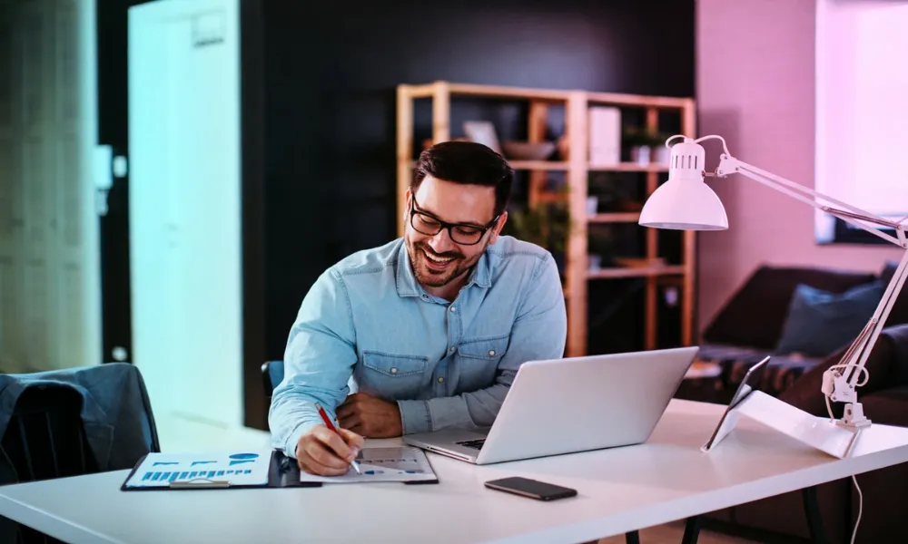 A man smiling while working on a laptop and taking notes at his desk.