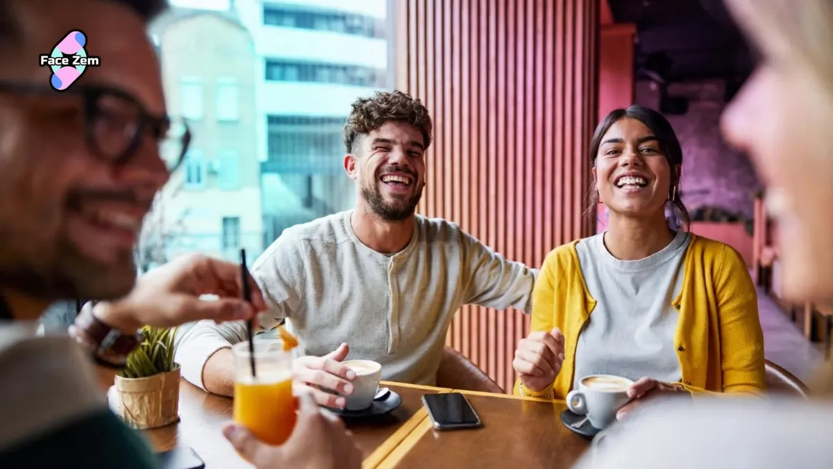 A group of young adults laughing and chatting together at a cafe table while drinking coffee.